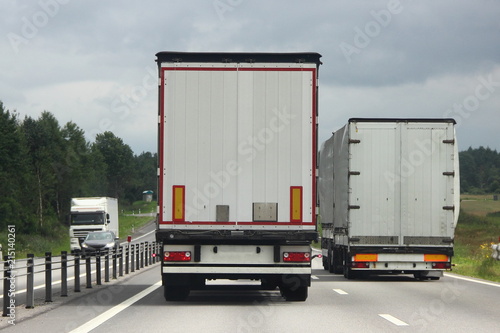 Logistics, international freight by road-two white trucks drive next to the two-lane asphalted country road in the summer day, rear view