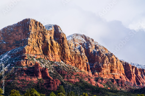 Rocky, rugged red sandstone hillside in Sedona, Arizona. Top is sprinkled with snow, cloudy sky is in the background. Sun is shining on one side; green of the Coconino forest is at the base. 