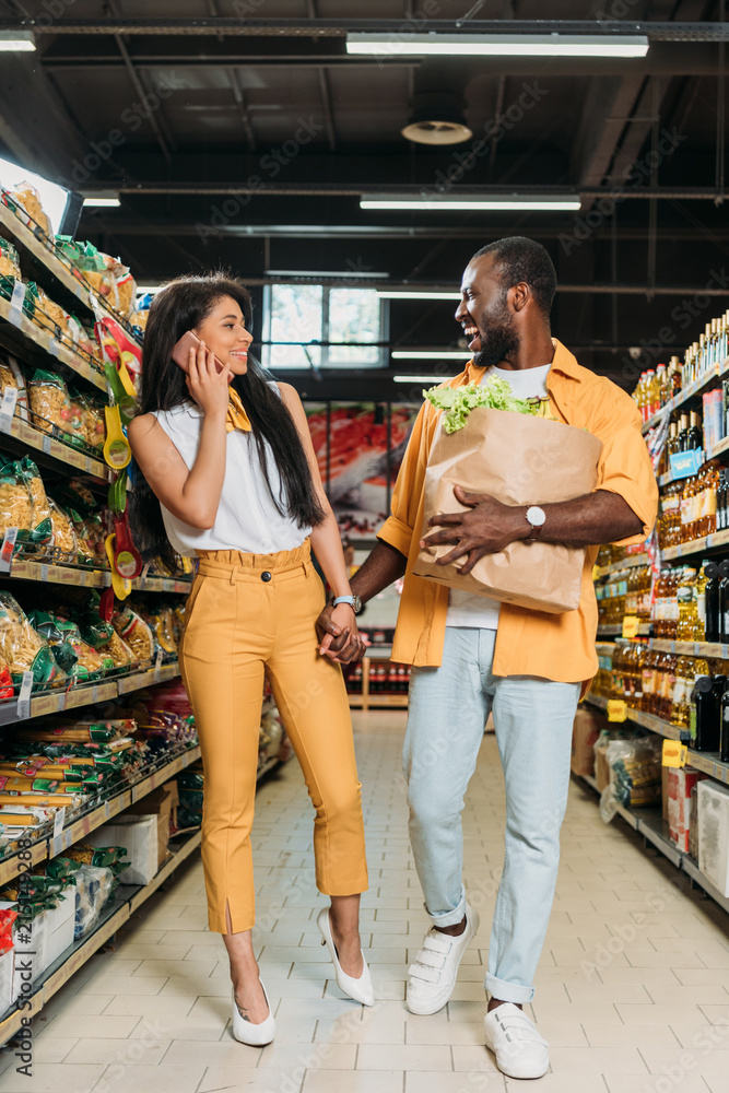 laughing african american man with paper bag holding hand of girlfriend while she talking on smartphone in supermarket