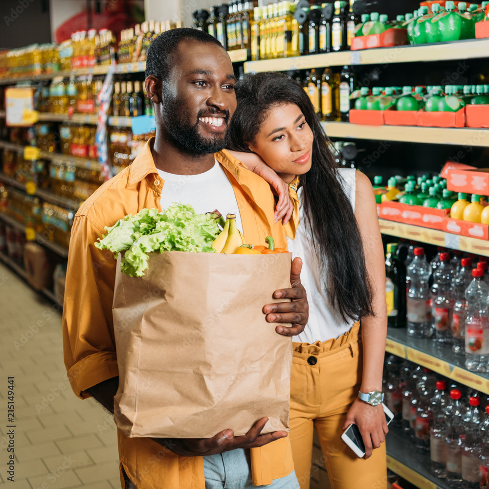 smiling african american couple with paper bag and smartphone in grocery store