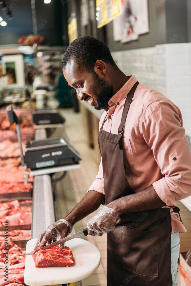 Φωτογραφία smiling african american male shop assistant in apron cutting steak of raw meat