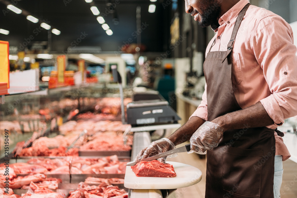 Φωτογραφία cropped image of african american male shop assistant in apron cutting raw meat