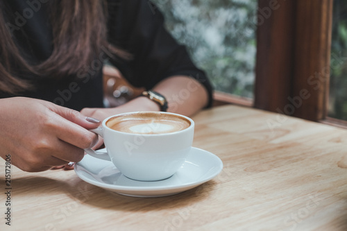Closeup image of a woman holding a white cup of hot coffee on wooden table in cafe