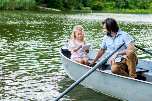 Happy father and daughter with tablet riding boat on lake at park