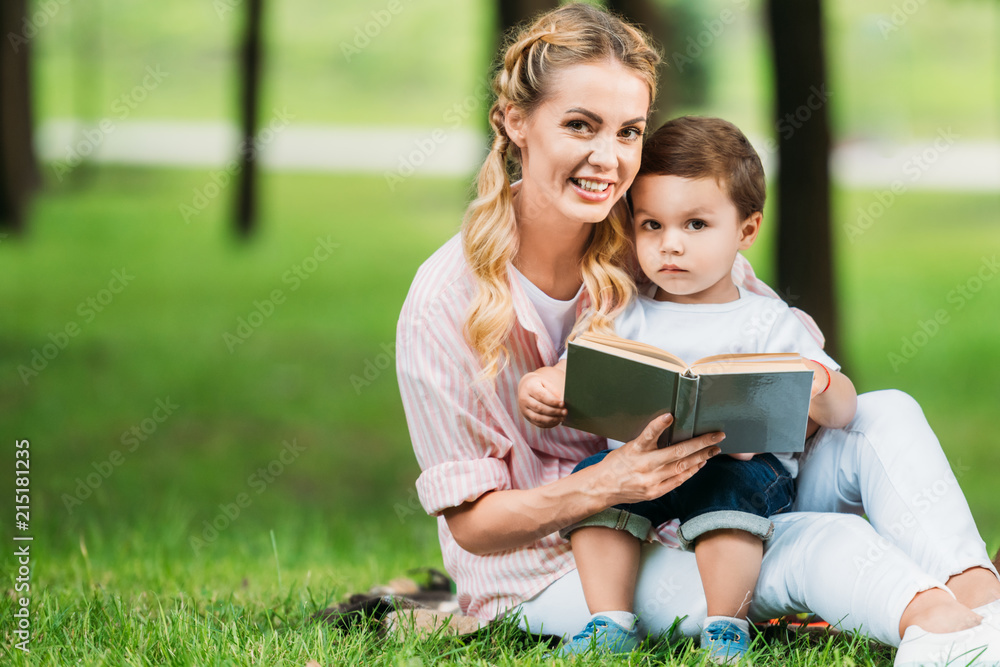 smiling mother and son sitting with book on grass at park and looking at camera smiling mother and son sitting with book on grass at park and looking at camera