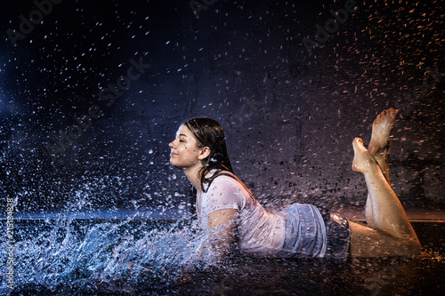 Girl with wet hair in the white shirt, water drops around and dark wall background illuminated by light