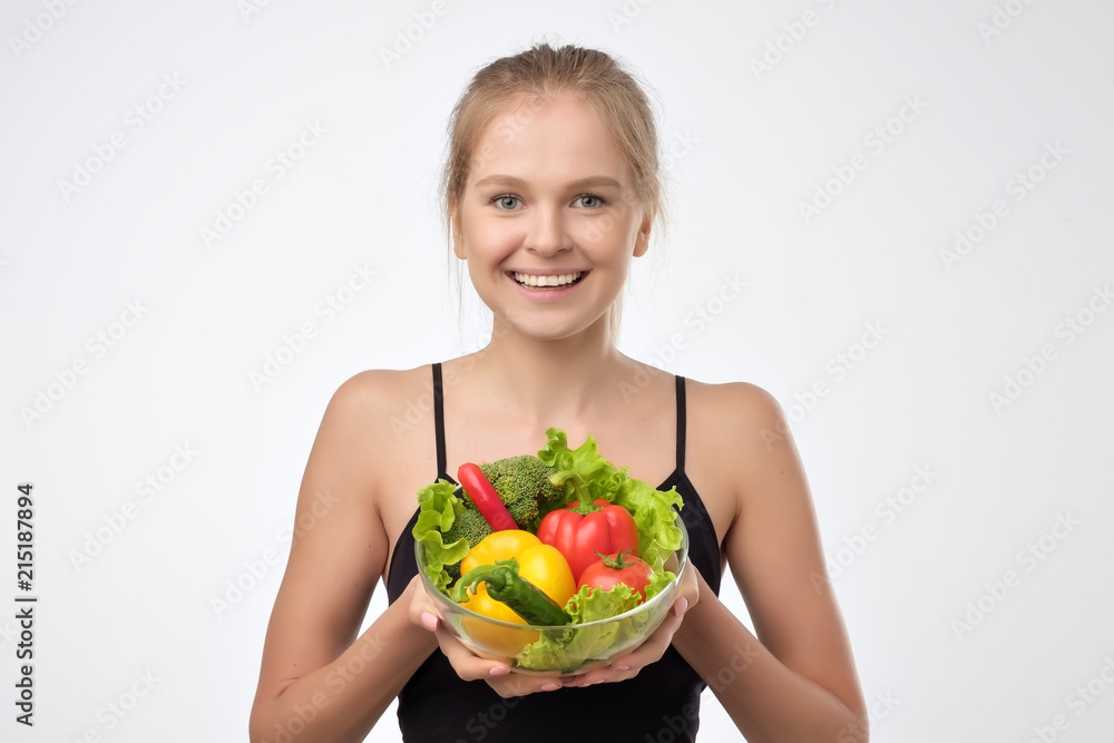 Smiling blonde young woman holding a pile of healthy fresh vegetables standing on white background. Concept of healthy diet eating Smiling blonde young woman holding a pile of healthy fresh vegetables standing on white background. Concept of healthy diet eating