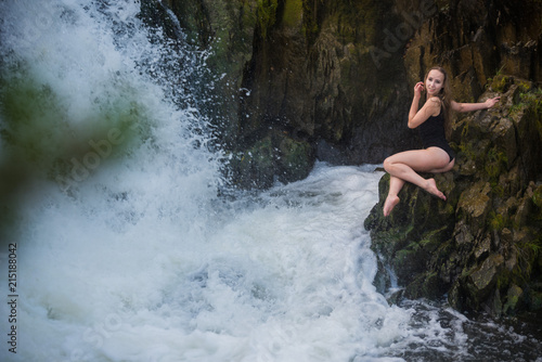 Young blond woman in a black swimwear sitting by the waterfall.  Young woman enjoying nature