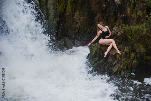 Young blond woman in a black swimwear sitting by the waterfall. Young woman enjoying nature