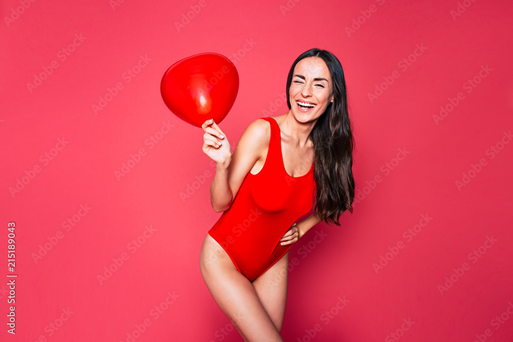 Happy Beautiful young smiling woman with long curly hair in red swimsuit posing over pink background with red balloon in hand Happy Beautiful young smiling woman with long curly hair in red swimsuit posing over pink background with red balloon in hand