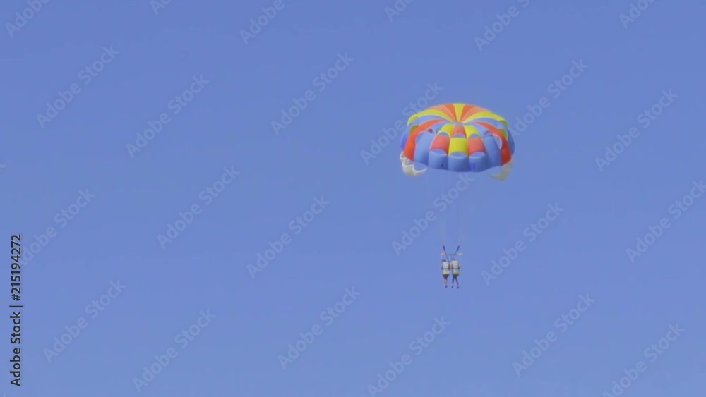 Two unrecognizable people are flying on a multi-colored parachute in the sky, which is tied to a motor boat. Parasailing in clear sunny weather Two unrecognizable people are flying on a multi-colored parachute in the sky, which is tied to a motor boat. Parasailing in clear sunny weather