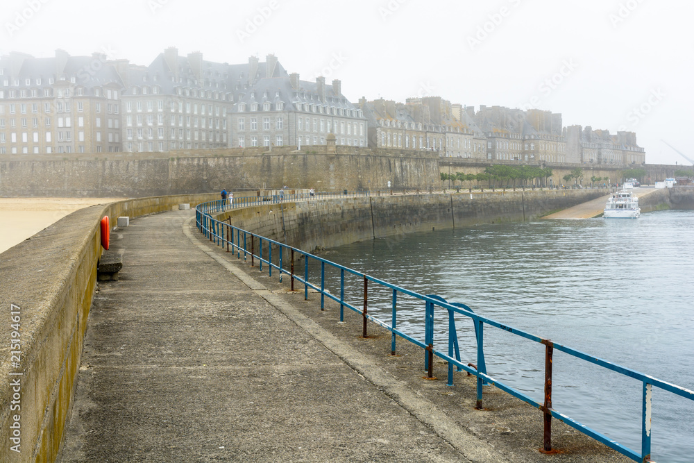 View from the breakwater of the walled city of Saint-Malo in Brittany, France, by a misty morning with the granite buildings sticking out above the ramparts and disappearing in the fog.