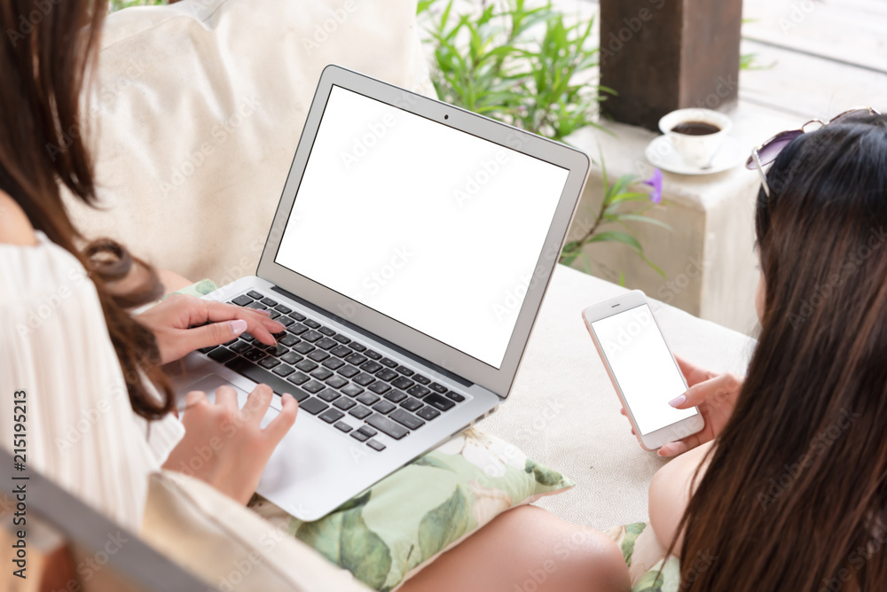 Two teenage women sit on big hammock use white blank screen laptop and smartphone while have coffee Two teenage women sit on big hammock use white blank screen laptop and smartphone while have coffee
