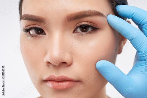 Hand in blue glove touching face of young Asian woman with pure skin on white background