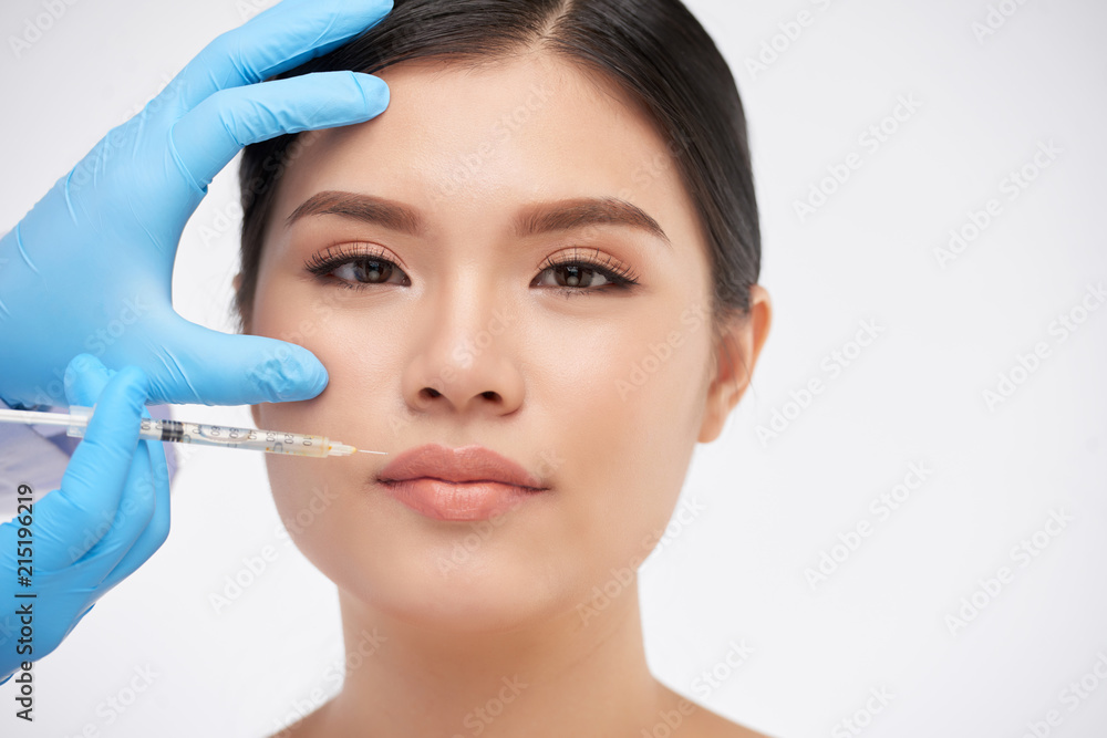 Crop hands in rubber gloves making beauty injection to pretty Asian woman looking at camera on white background Crop hands in rubber gloves making beauty injection to pretty Asian woman looking at camera on white background