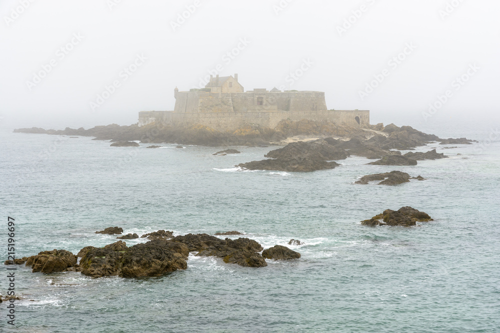 The Fort National built by french military architect Vauban on a tidal island, seen from the city of Saint-Malo, France, disappearing in the fog with rocks in the sea in the foreground.