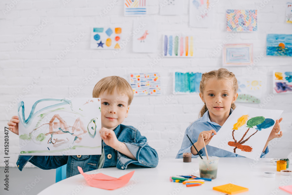 Fototapeta premium portrait of cute kids showing drawings in hands at table in classroom