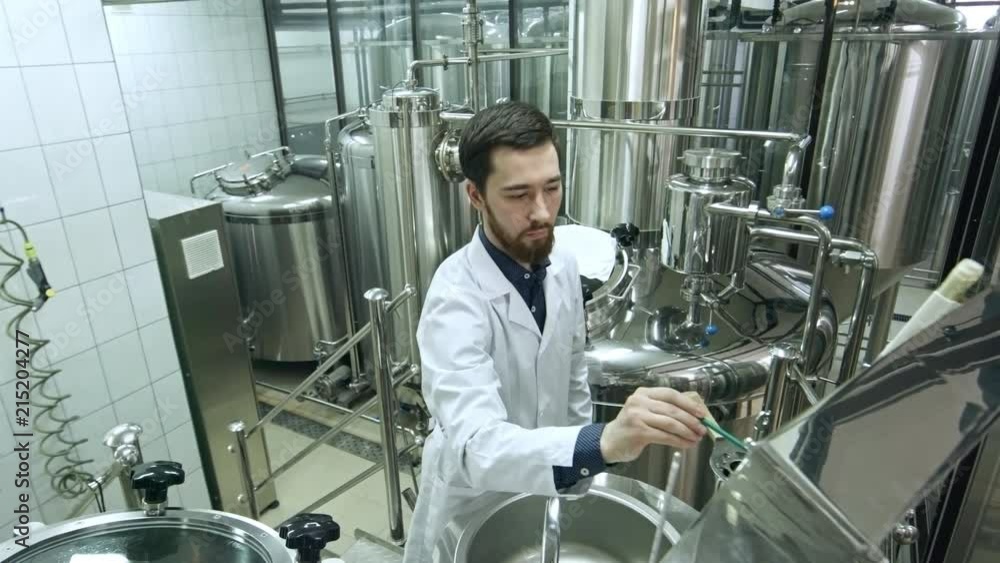 Professional brewery worker in lab coat working with sensor display while setting up modern brewing machine and checking boiling beer in vessel Professional brewery worker in lab coat working with sensor display while setting up modern brewing machine and checking boiling beer in vessel