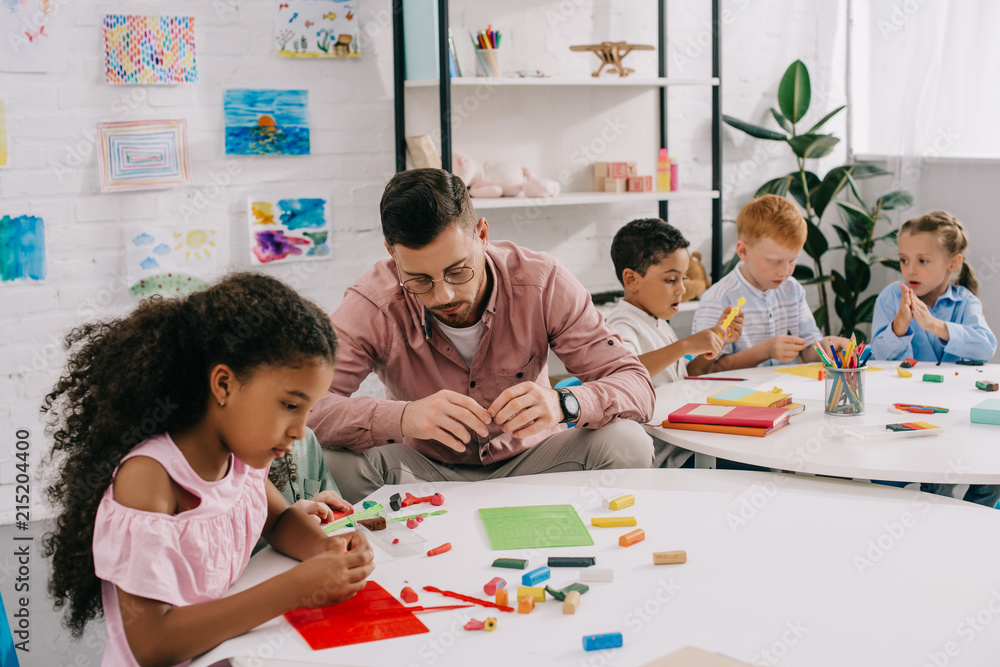 teacher and multiethnic kids sculpturing figures with colorful plasticine at table in classroom