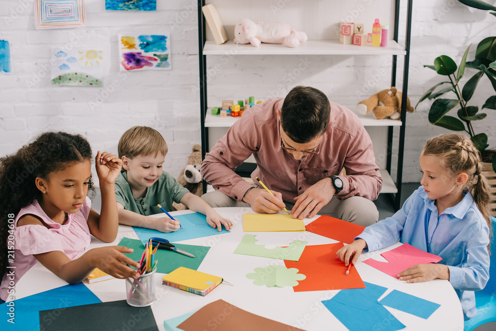 Fototapeta premium multiethnic preschoolers and teacher making paper applique at table in classroom