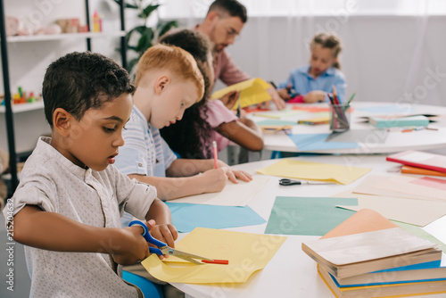 selective focus of teacher and interracial preschoolers cutting colorful papers with scissors in classroom
