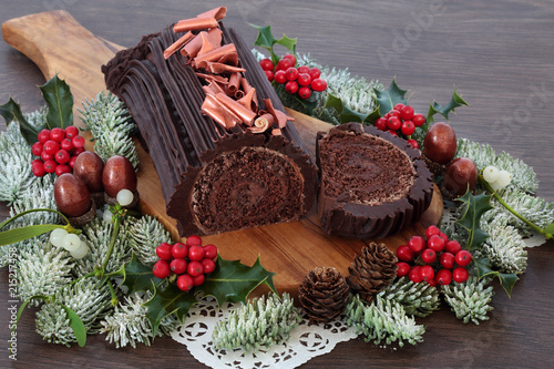 Chocolate yule log Christmas cake with winter flora of snow covered fir, holly, acorns and mistletoe on rustic oak wood background. Festive food  for the holiday season.