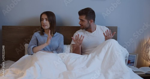 Caucasian just married man and woman having a quarrel and being offended at each other while sitting in the bed under the blanket in the evening. photo