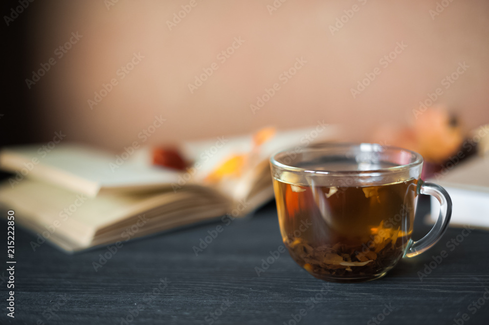 Fototapeta premium Autumn concept. Tea in a transparent mug, unfolded book, yellow and red autumn leaves and spruce branches, cones and chestnuts on a dark background