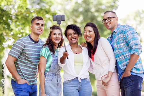 people, friendship and international concept - happy smiling young woman and group of happy friends taking picture by selfie stick in park