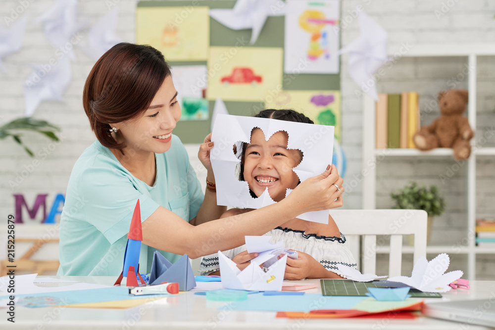 Fototapeta premium Asian teacher putting paper with flower cut-out to face of little girl in art class