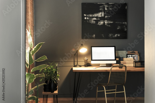 Gold chair standing by the wooden desk with lamp, notebooks and mockup computer in real photo of dark living room interior with modern poster on the wall © Photographee.eu