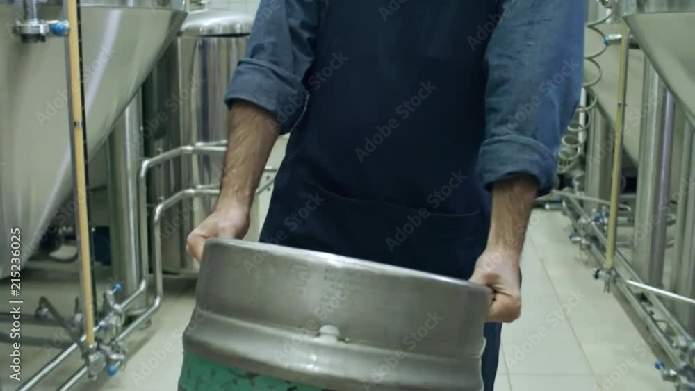 Tilt down of bearded worker in apron carrying heavy beer keg and walking through brewery plant Tilt down of bearded worker in apron carrying heavy beer keg and walking through brewery plant