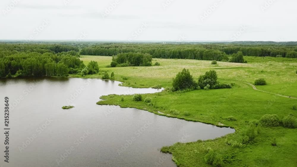 Flying over the lake, green meadows and forest on cloudy summer day Flying over the lake, green meadows and forest on cloudy summer day