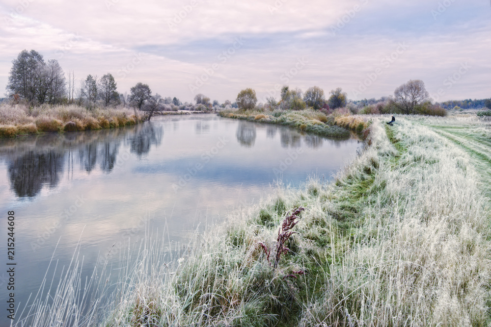 Beautiful Autumn Belarusian Landscape On The Topic Of Fishing: Lone Fisherman Sits On The Grassy Bank Of The River, Covered With Hoarfrost. On Fishing: Waiting For A Bite. Cold Autumn Morning, Belarus