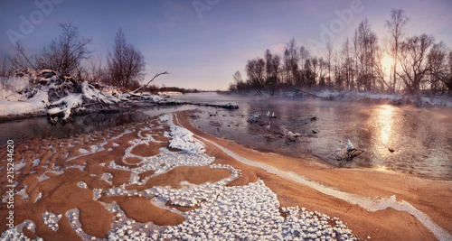 Obraz na płótnie A Beautiful Winter Morning Landscape: The Sun Rising Over The Forest And The Sandy Shallows Of The River, Covered With Frost Crystals. Belarusian Winter Panoramic Landscape With Sun, River And Forest w salonie