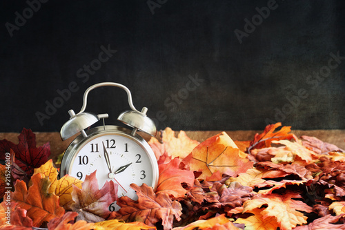 Alarm clock in colorful autumn leaves against a dark background with shallow depth of field. Daylight savings time concept.