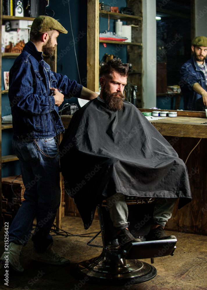 Barber preparing hair clipper for bearded man, barbershop background. Barber with clipper and brutal bearded client. Hipster client covered with cape waiting for haircut. Hipster lifestyle concept