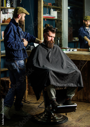 Barber preparing hair clipper for bearded man, barbershop background. Barber with clipper and brutal bearded client. Hipster client covered with cape waiting for haircut. Hipster lifestyle concept © be free