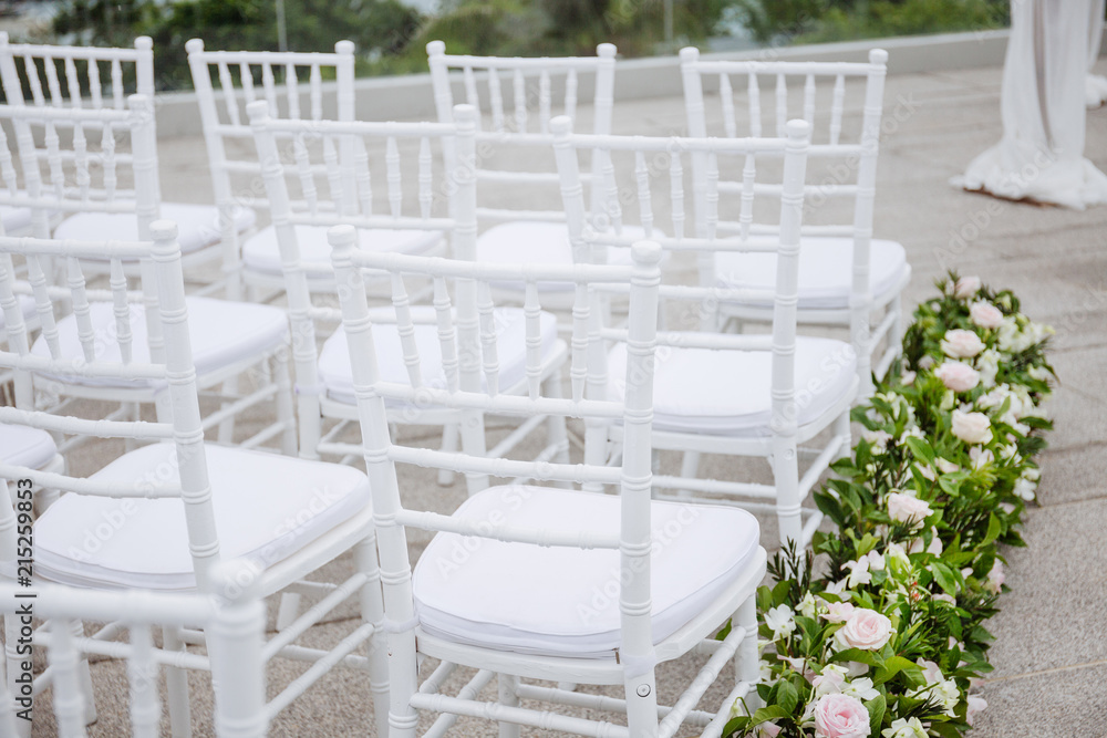 Close-up back side of white chiavari chairs arrangement for modern wedding ceremony, white and pink roses and green leaf decoration on the aisle walk way