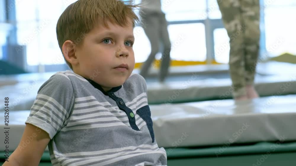 Medium shot of tired or bored boy of elementary school age sitting and relaxing on trampoline in indoor play center, other children jumping in the background Medium shot of tired or bored boy of elementary school age sitting and relaxing on trampoline in indoor play center, other children jumping in the background