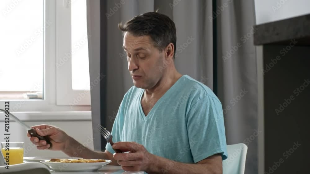 Medium shot of middle-aged man sitting at table in kitchen and talking to someone before eating scrambled eggs for breakfast Medium shot of middle-aged man sitting at table in kitchen and talking to someone before eating scrambled eggs for breakfast