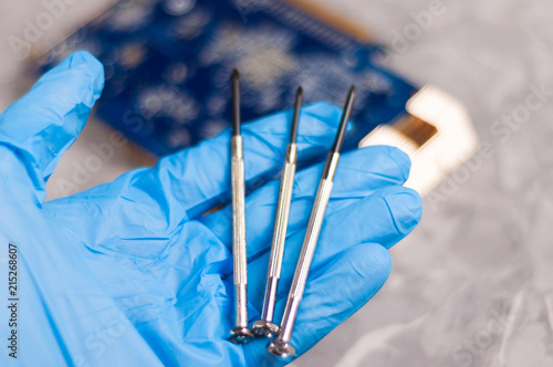 Mens hand in blue rubber glove holds three small metal screwdrivers on background of pc graphics card with copper radiator on old gray concrete floor