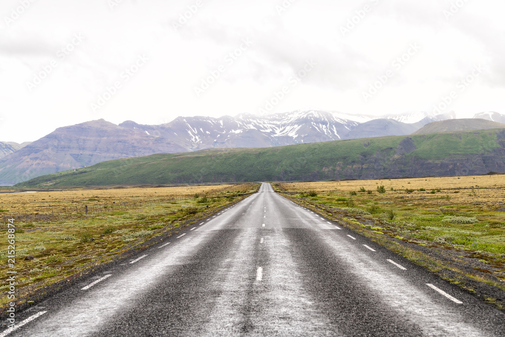 View of Iceland road route 1 center, middle with vanishing point lines, snow covered capped mountains, cliffs, green grass, moss in summer, cloudy mist sky