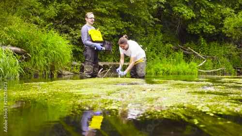 Man and woman scientist environmentalist standing in a river. Woman taking sample of water and pouring it into the test tube. Man holding toolbox photo
