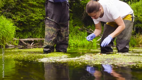 Man and woman scientist environmentalist standing in a river. Woman taking sample of water plant and putting it in a toolbox. Man holding toolbox