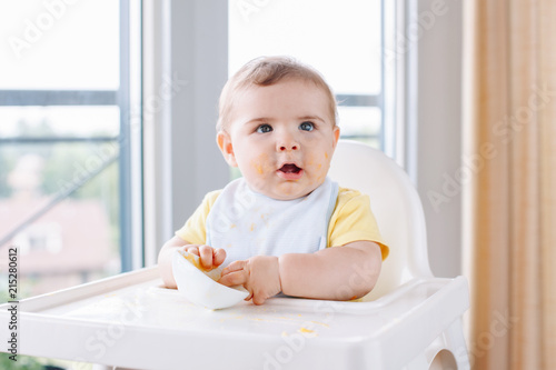 Portrait of cute adorable Caucasian child boy with dirty messy face sitting in high chair eating apple puree with fingers. Everyday home childhood lifestyle. Infant trying supplementary baby food