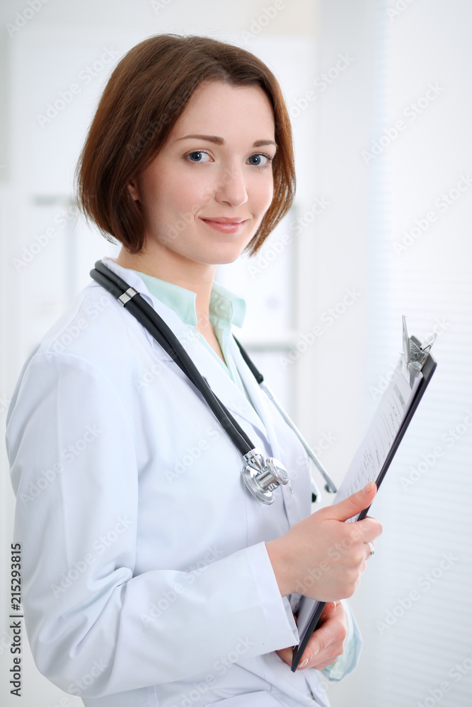 Young brunette female doctor standing with clipboard and smiling in hospital Young brunette female doctor standing with clipboard and smiling in hospital