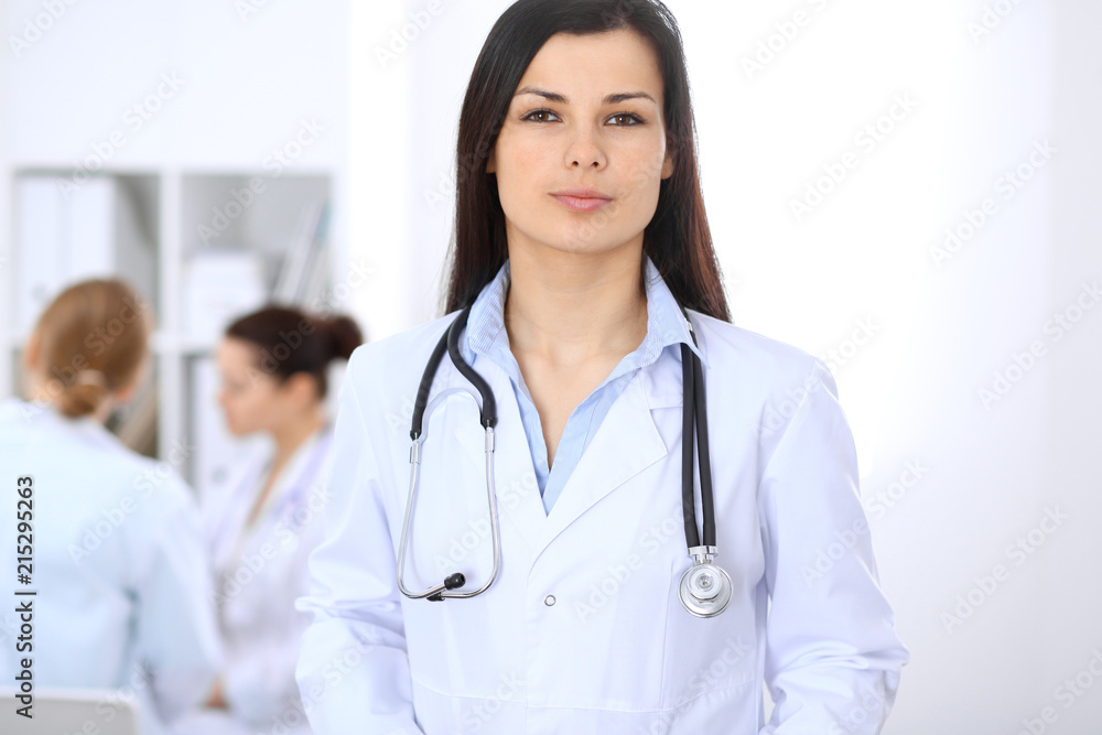 Brunette female doctor on the background of colleagues talking to each other in hospital. Medicine and health care concept Brunette female doctor on the background of colleagues talking to each other in hospital. Medicine and health care concept
