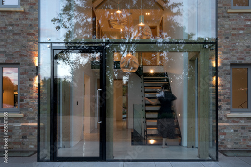 Evening view of the glass entrance to a modern house with lights on and a woman ascending the stairs.