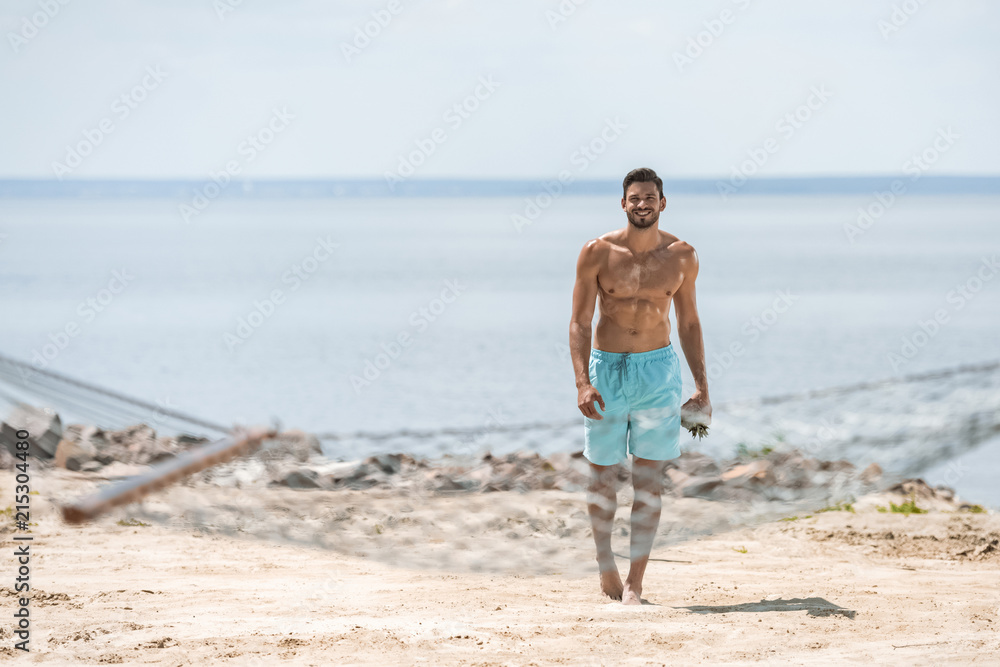 handsome shirtless man walking on beach, with hammock on foreground handsome shirtless man walking on beach, with hammock on foreground
